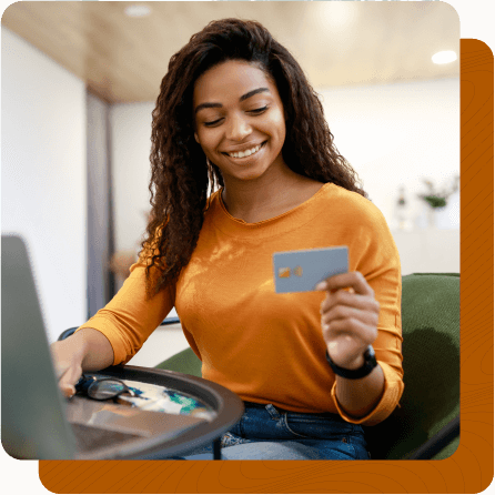 Happy woman sitting in chair at table looking at credit card in one hand and using laptop in the other.