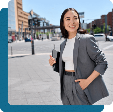 Business woman in dress suit walking in company square with large buildings behind her holding tablet in hand.