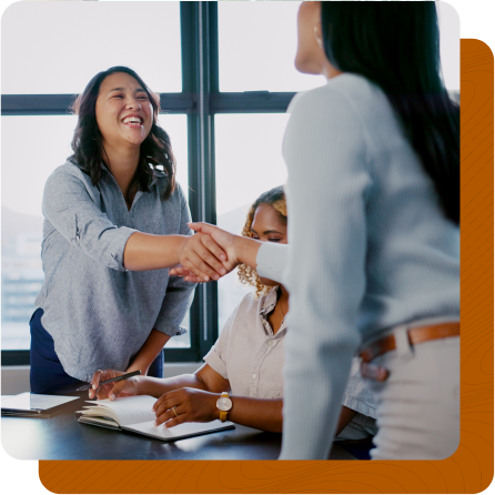 Two women shaking hands across table while other woman sits down in between them.