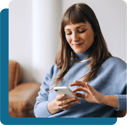 Woman with bangs and blue sweaters smiling while looking at and using cell phone.