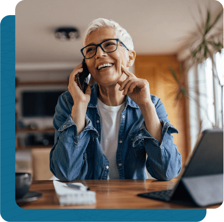 Woman sitting at table in living room talking on the phone with tablet and notebook in front of her.