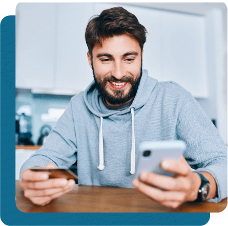 Young man working at table in kitchen with credit card in one hand and phone in the other.