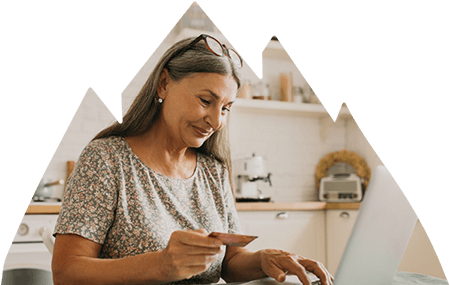 Older woman sitting at kitchen table working on laptop while holding credit card in hand.