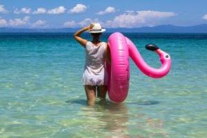 Stylish pretty women on summer vacation on tropical beach holding inflatable pink flamingo enjoying in her freedom and incredible sea view. Travel destination. Aegean sea, Greece.
