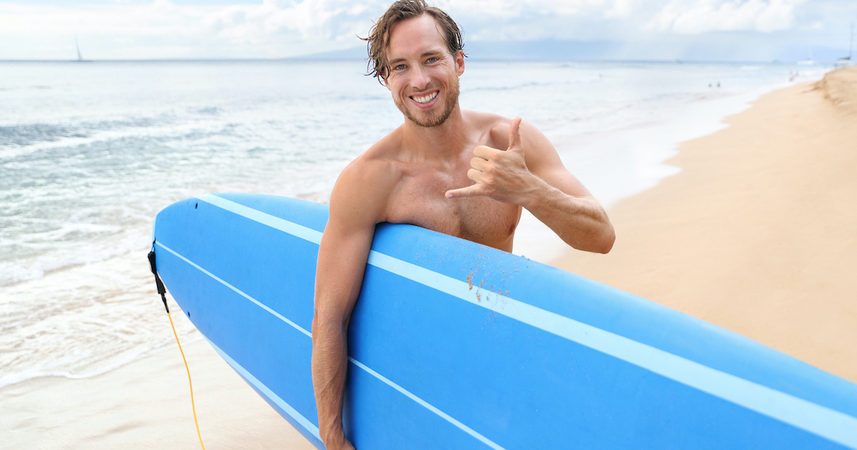 Surfer guy happy with surf surfing smiling doing hawaiian shaka hand sign for fun during surf session in ocean waves on beach vacation. Surfing travel destination. Friendly greeting in surfer culture.